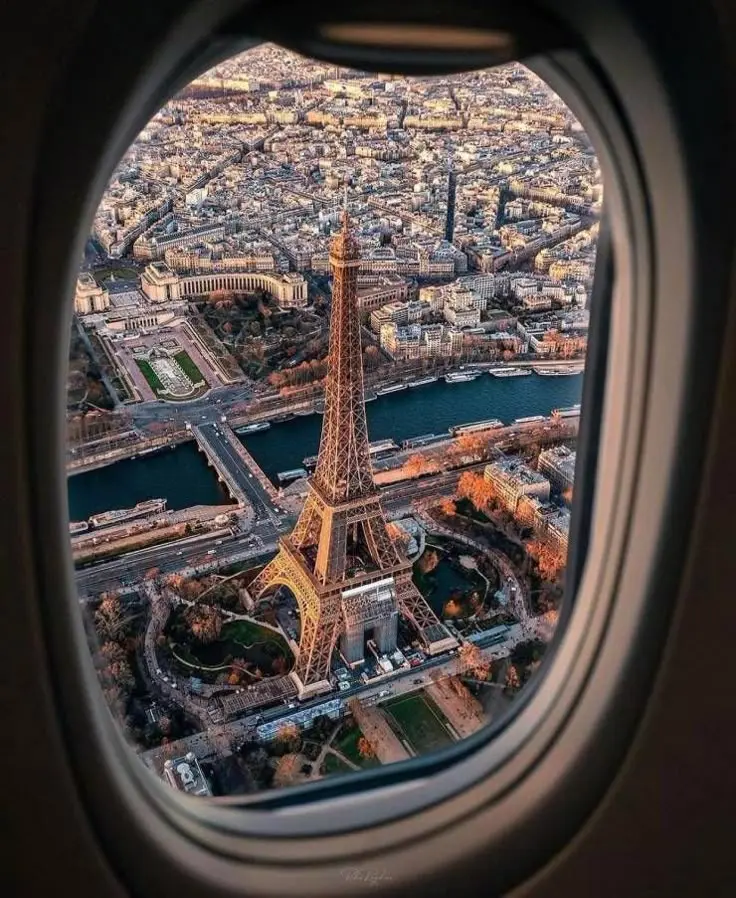 Aerial view of the Eiffel Tower at sunset, seen through an airplane window. The Seine River and surrounding Parisian architecture are visible, bathed in golden light.