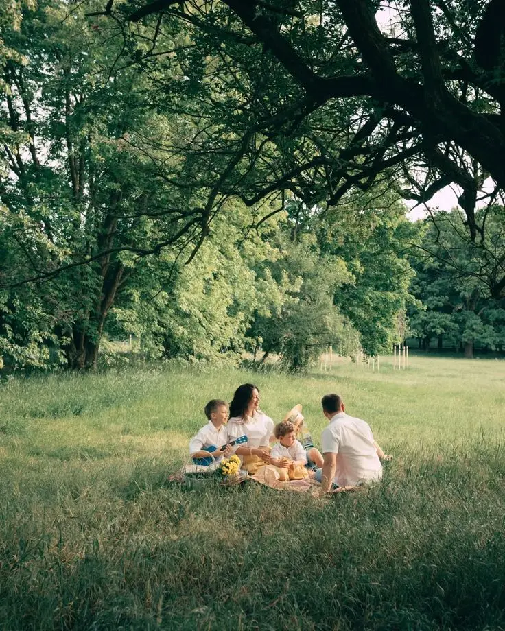 A family picnics under a tree in a sunlit meadow. They sit on a blanket, sharing snacks and laughter, surrounded by lush, green trees, conveying joy.