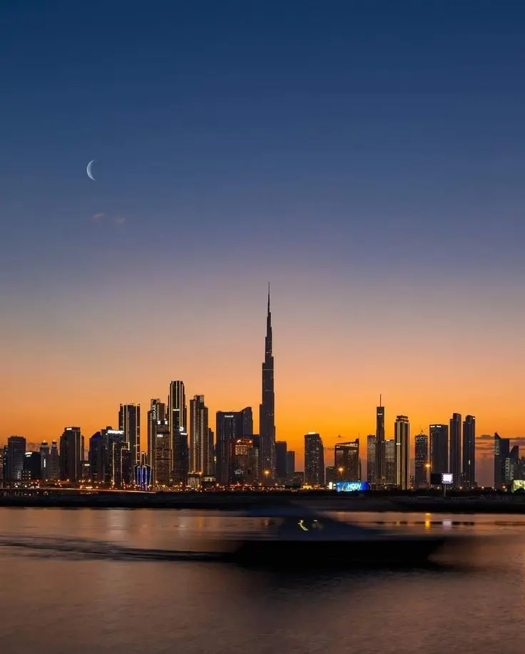 Dubai city skyline at sunset with the Burj Khalifa in the center, a crescent moon in the sky, and a blurred boat on the water in the foreground.