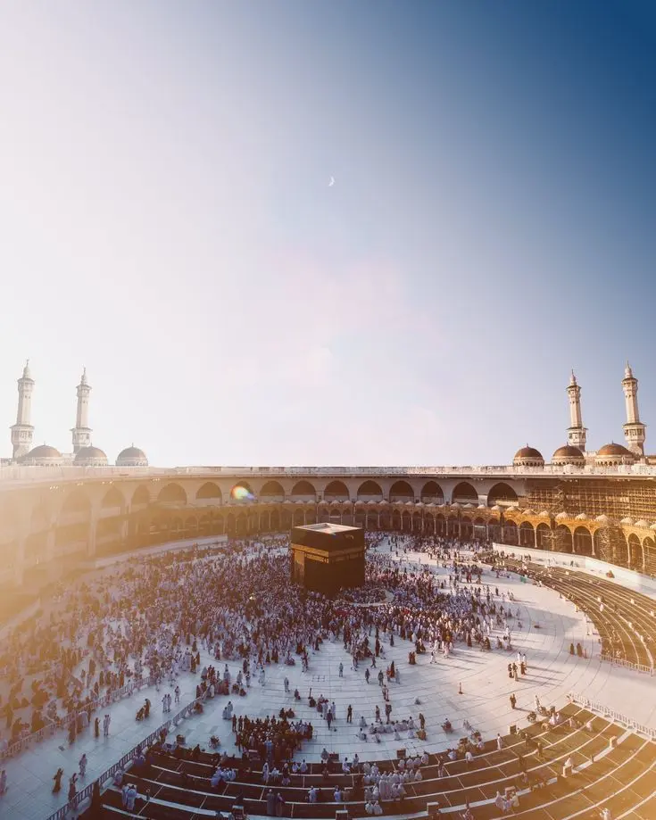 Aerial view of the Kaaba surrounded by pilgrims at the Grand Mosque in Mecca. Minarets stand tall against a clear, light blue sky at sunrise.